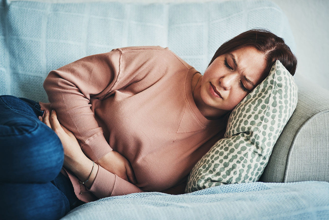 A woman holds her abdomen in pain while laying down on a sofa at home