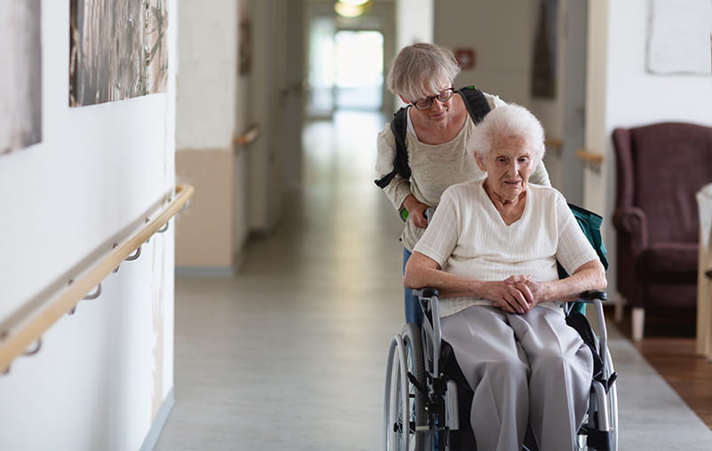 A woman pushes an older woman in a wheelchair down a hallway.