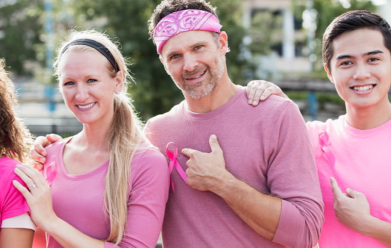 Two smiling men and a woman wear pink shirts for breast cancer awareness.