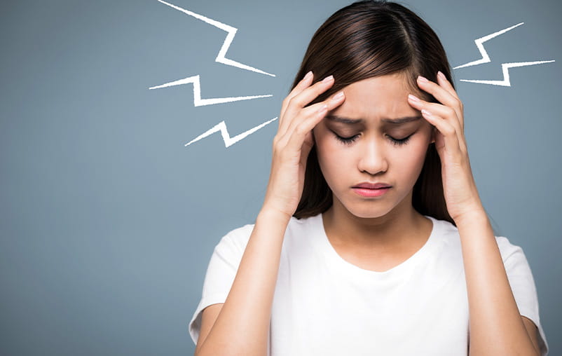 A teenage girl holds her hands to her temples in pain. Illustrated zigzags shoot out from the sides of her head.