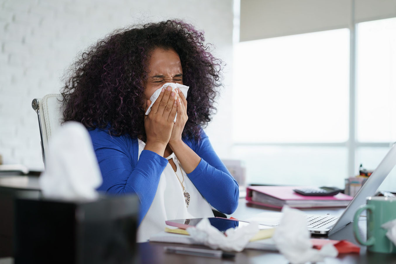 A woman blows her nose while sitting at a desk
