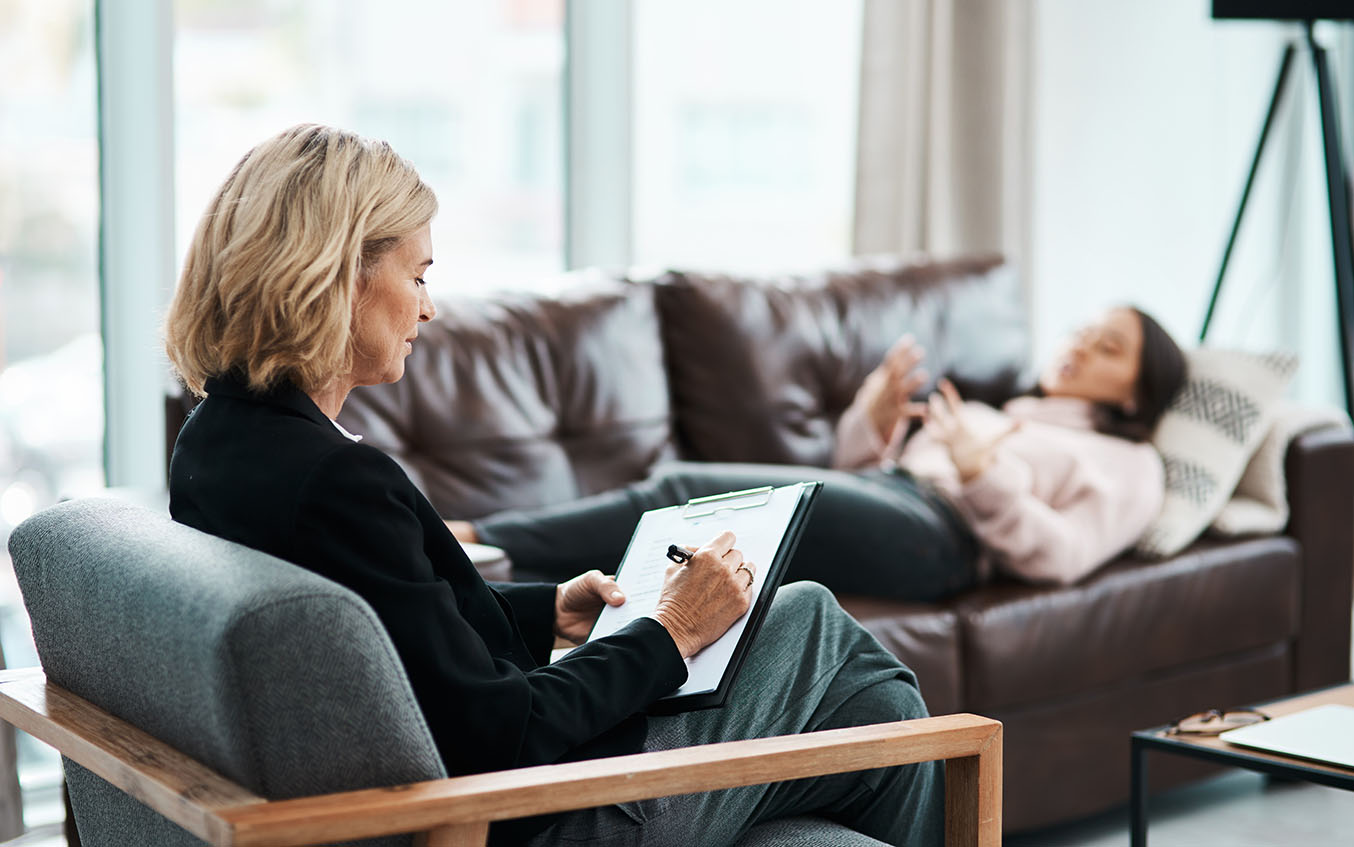 A woman reclines on a couch while talking to a therapist, who is sitting in a chair taking notes.