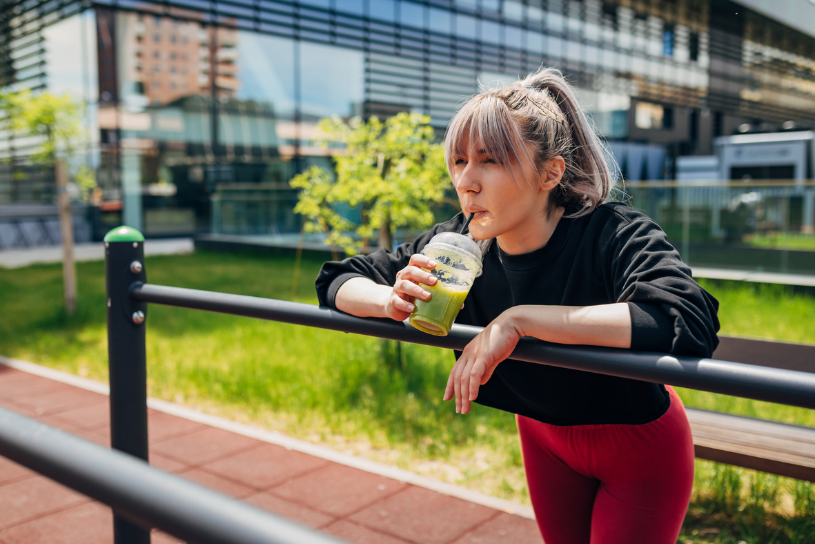 Woman in gym gear leaning over a metal fence drinking a smoothie.